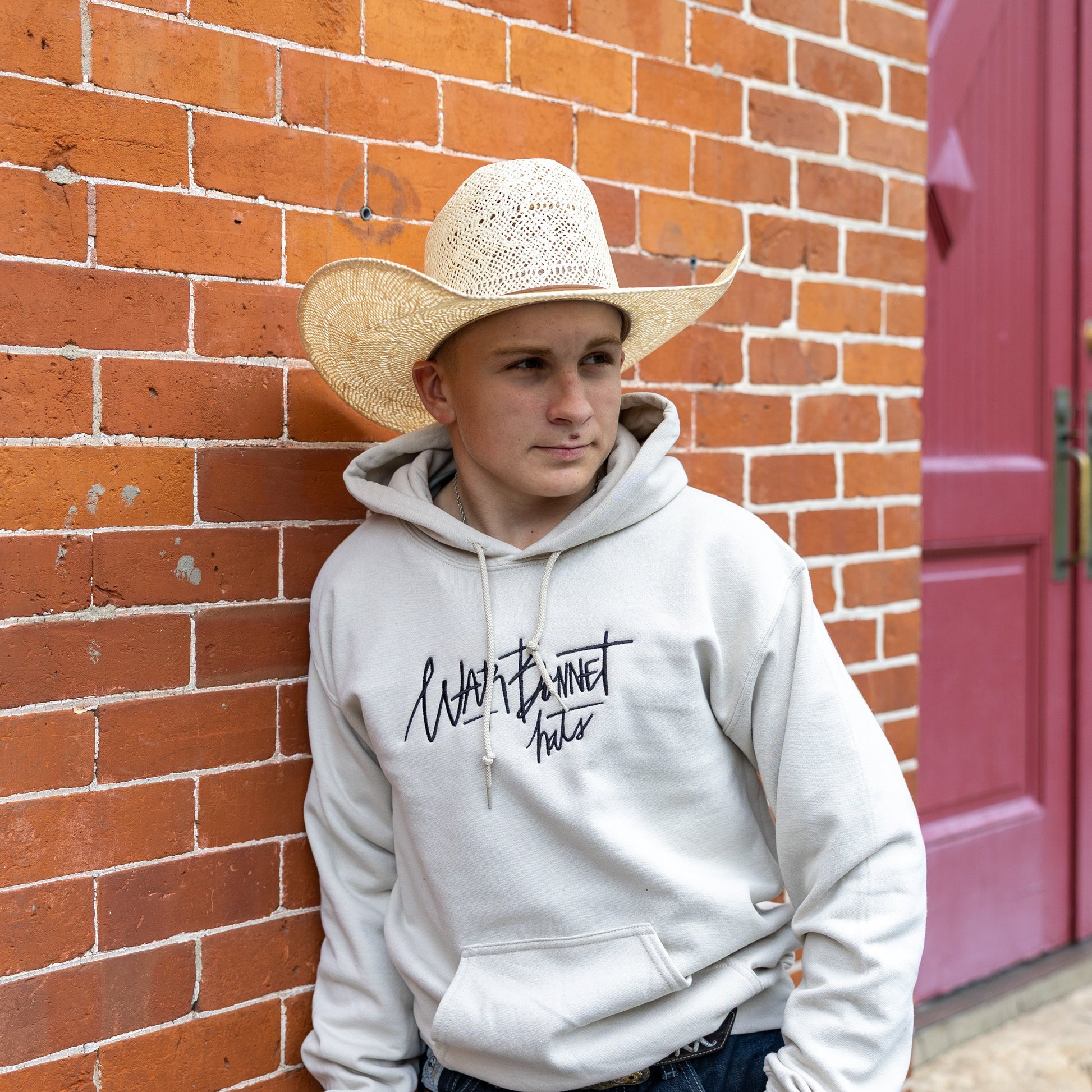 A young man wearing a cream cowboy hat and a light gray hoodie with 'Warbonnet Hats' printed on the chest, leaning against a red brick wall next to a red door.
