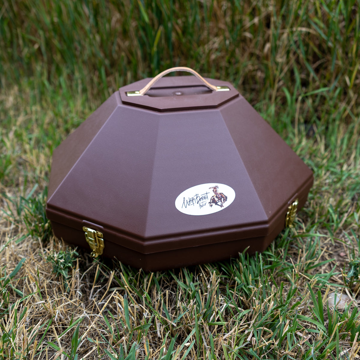 A brown octagonal hat can with a top handle, resting on grass. It features a white oval sticker on the front with a logo and the text 'Warbonnet Hats'.
Large enough to fit a 5 inch brim hat. 