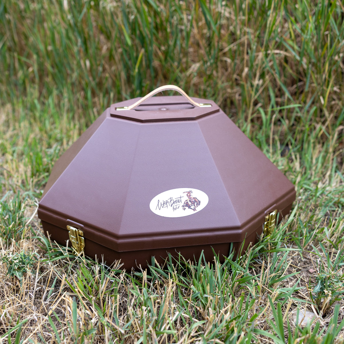 A brown octagonal hat can with a top handle, resting on grass. It features a white oval sticker on the front with a logo and the text 'Warbonnet Hats'.
Large enough to fit a 5 inch brim hat. 
