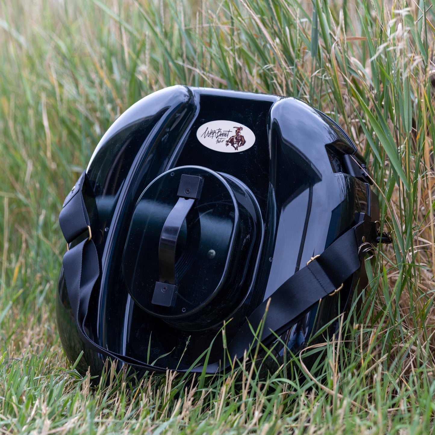 A glossy black hat carrier resting on green and dry grass outdoors. The hat can features a circular white and brown logo sticker on the front. It is a better hat can, with a strap for easy transport.