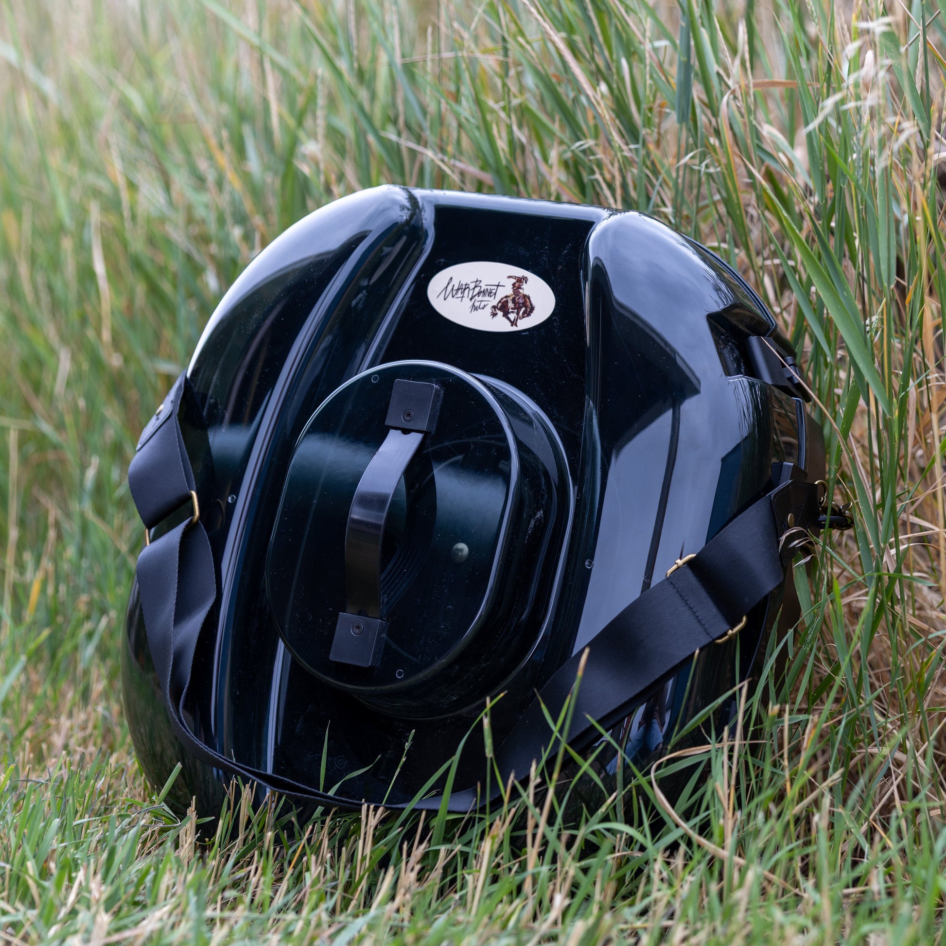 A glossy black hat carrier resting on green and dry grass outdoors. The hat can features a circular white and brown logo sticker on the front. It is a better hat can, with a strap for easy transport.