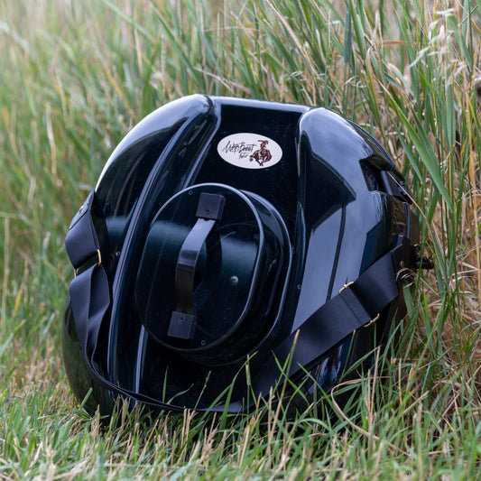 A glossy black hat carrier resting on green and dry grass outdoors. The hat can features a circular white and brown logo sticker on the front. It is a better hat can, with a strap for easy transport.